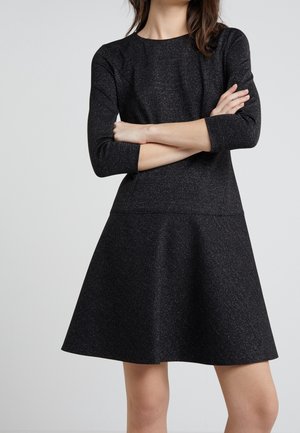 Mujer con un vestido negro de manga larga hasta la rodilla con un leve brillo, brazos cruzados, sobre un fondo liso de color gris claro.