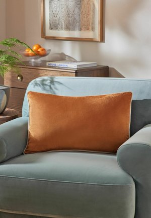 Hellblauer Sessel mit großem rechteckigem Kissen in verbranntem Orange, Holzsideboard mit Buch und Schale mit Orangen sowie gerahmte abstrakte Wandkunst.