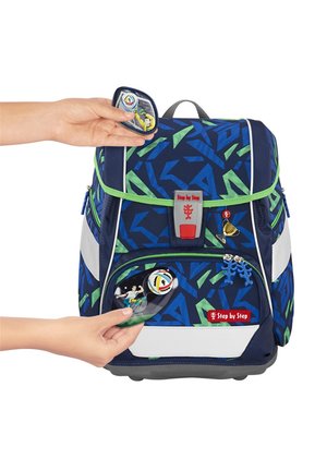 Blauer und grüner Rucksack mit gemustertem Design und reflektierenden Akzenten, rotem und grauem Logo sowie abnehmbaren Patches. Hand befestigt ein Patch an der Vordertasche.