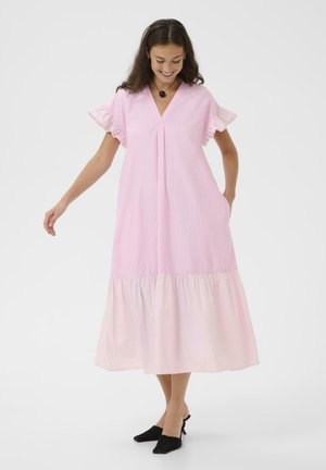 Kobieta w różowej sukience midi z falbankami na rękawach i brzegu, ubrana w czarne szpilki i okrągły czarny naszyjnik, uśmiechająca się i patrząca w dół.