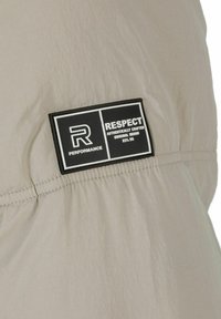 Lysegrå stof med en krøllet tekstur har et sort rektangulært label med hvid tekst, der står "RESPEKT" og "YDEEVNE."