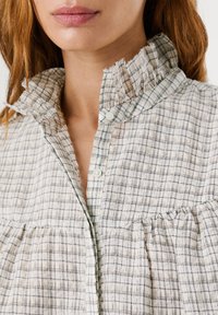 Femme portant un chemisier blanc avec un fin motif en quadrillage noir et un col montant à volants, partiellement boutonné au niveau du cou.