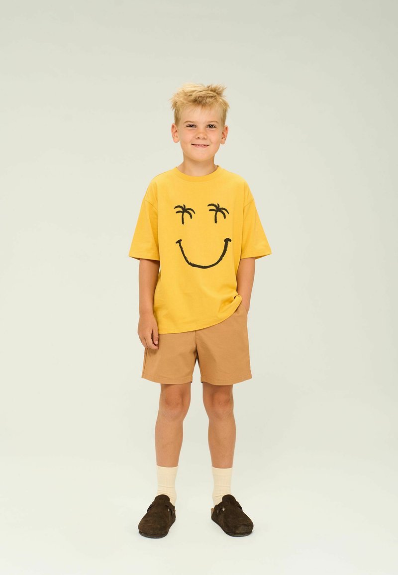 Gul T-shirt med sort smiley-ansigt design, parret med tan shorts. Båret med brune slip-on sko og lyse sokker.