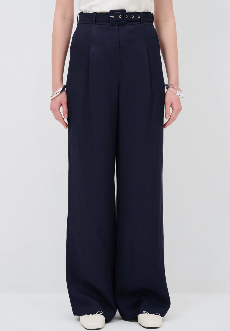 Pantalons larges marine à taille haute avec ceinture, comportant des plis et une texture de tissu lisse. Associés à des chaussures de couleur claire.