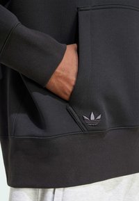 Svart sweatshirt med en strukturerad tyg, kängurufickor och en subtil grå Adidas-logga på fållen. Har ribbade cuffs och midja.
