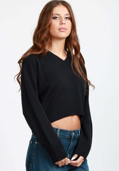 Giovane donna con lunghi capelli castani ondulati, che indossa un maglione nero corto e jeans blu a vita alta, in piedi contro uno sfondo bianco.