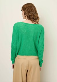 Groene, loszittende trui van een zachte, gestructureerde stof. Heeft lange mouwen en een wijd decolleté, gecombineerd met beige broeken.