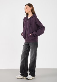 Corta-vento roxo escuro com fecho, com dois bolsos frontais e ferragens prateadas, combinado com calças largas pretas e ténis cinzentos.