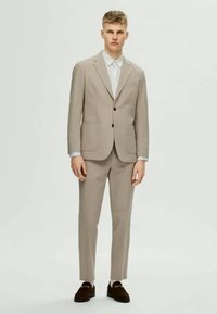 Beżowy garnitur z wyciętymi klapami i dwiema przednimi kieszeniami, noszony na białej koszuli. Ciemne mokasyny dopełniają stylizację. Cały zestaw ma nowoczesny krój.