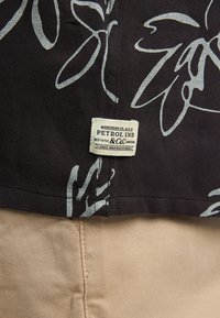 Czarna tkanina z białym wzorem kwiatowym; widoczna metka z napisem "PETROL IND" znajduje się blisko brzegu, obok beżowego materiału poniżej.