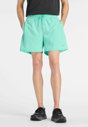 Mann trägt türkisfarbene Sportshorts, schwarze Turnschuhe und ein schwarzes Shirt, steht mit den Händen in den Taschen vor einem weißen Hintergrund.