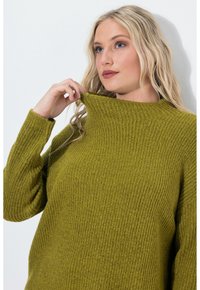 Maglione lavorato a maglia verde oliva con collo alto, texture a coste e vestibilità ampia. La modella sta sollevando delicatamente il collo.