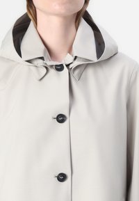 Impermeabile grigio chiaro con cappuccio, caratterizzato da una trama liscia, bottoni neri e un colletto con un design arrotondato.