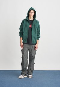 Sweatshirt verde com fecho à frente e bolso tipo canguru, combinado com calças cargo cinzas e ténis cinzentos. A sweatshirt tem um logótipo no peito.