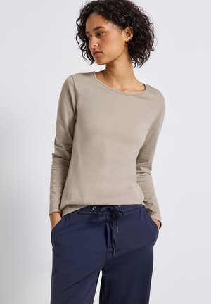 Femme avec des cheveux bouclés et foncés portant une chemise beige à manches longues et un pantalon bleu marine avec cordon de serrage, les mains dans les poches, regardant sur le côté.