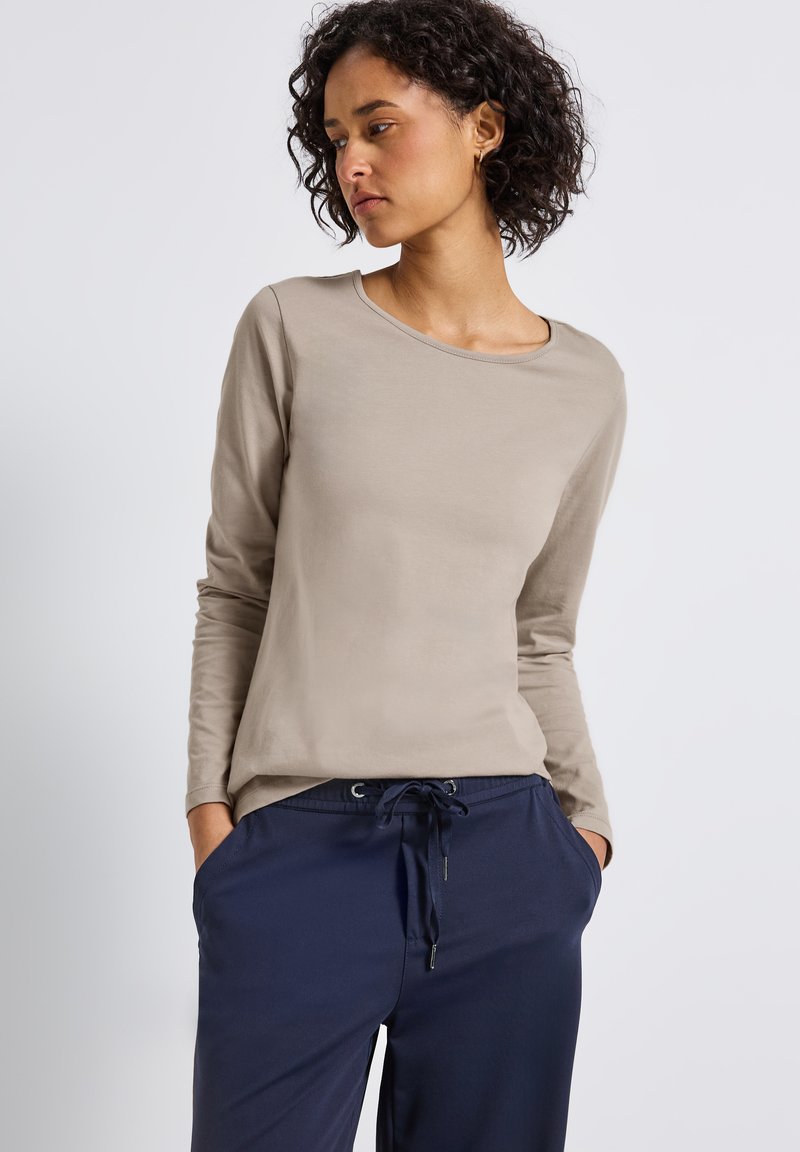 Donna con cappelli ricci scuri, che indossa una camicia a maniche lunghe beige e pantaloni con coulisse blu navy, con le mani nelle tasche, che guarda di lato.