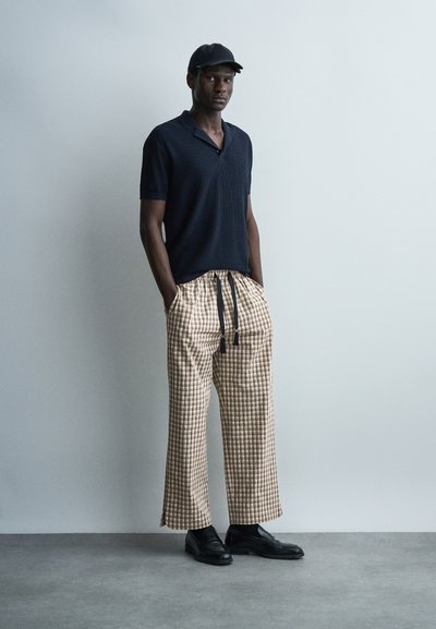 Homme debout, mains dans les poches, portant une casquette noire, une chemise à manches courtes sombre, un pantalon large beige à carreaux et des chaussures noires sur un fond uni.