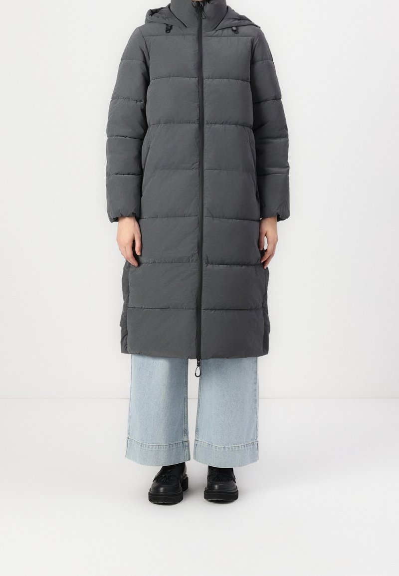 Grijs gewatteerd jas met een capuchon, lange mouwen en een rits aan de voorkant. Heeft rechthoekige stiknaden en is tot halverwege de kuit. Wordt gedragen over wijde jeans.