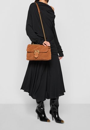 Femme en robe noire à une épaule, portant des bottines noires à talons et tenant un sac à bandoulière en suède marron avec une chaîne et une fermeture dorées.