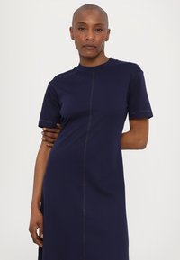 Marineblaues, kurzärmliges Kleid aus weichem Stoff mit geradem Schnitt, einem zentralen Naht und kontrastierenden Nähdetails.