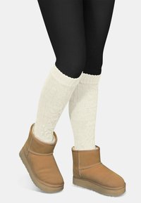 Braune Knöchelstiefel aus weichem Wildleder mit abgerundeter Zehenform und heller Sohle, kombiniert mit weißen, kabelgestrickten Beinstulpen über schwarzen Leggings.