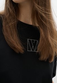 Melnā T-kreklā ir rhinestone izšūta ģeometriska logo priekšpusē. Tam ir gluda faktūra, apaļa kakla izgriezums un brīvi pieguļošs griezums.