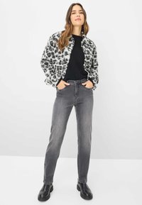 Donna che indossa un top nero, jeans grigi, stivali neri e un giubbotto con stampa leopardata bianca e grigia, in piedi con le mani in tasca.