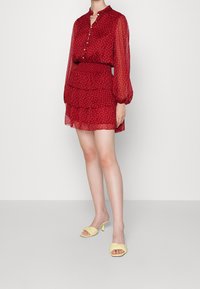 Punainen läpikuultava mekko, jossa on kukkakuosi, pitkät puffihihat ja kerroksellinen helma; yhdistetty keltaisiin korkokenkiin. Kevyt ja valuva kangas.