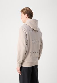Beige kapucnis pulóver, elülső kenguru zsebbel, bordás mandzsettákkal, és a hátán a "CALVIN KLEIN JEANS" felirat fekete betűkkel.