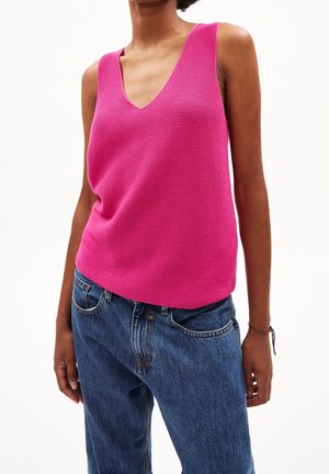 Femme portant un top en tricot sans manches rose vif avec un col en V et un jean denim bleu, debout devant un fond blanc uni.