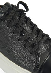 Schwarzer Sneaker mit strukturiertem Lederobermaterial, schwarzen Schnürsenkeln, Metallösen und einer weißen Gummisohle. Nahaufnahme, die Details und die Textur des Schuhs zeigt.