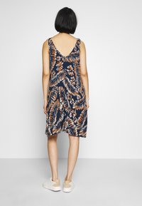 Robe bleu marine avec un motif floral tropical en orange et blanc. Design sans manches avec un dos en V et un ourlet fluide, portée avec des chaussures blanches.