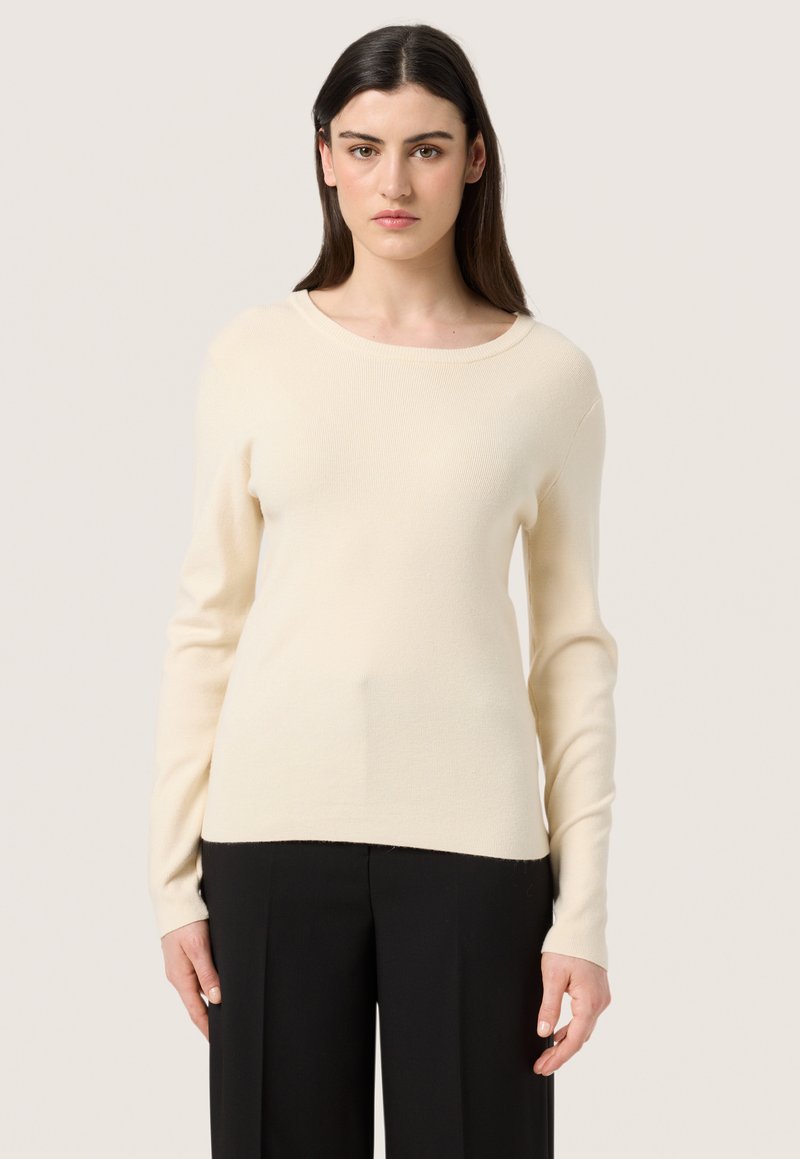 Cremfarbener gerippter Pullover mit Rundhalsausschnitt und langen Ärmeln, aus weichem Stoff, kombiniert mit schwarzen, maßgeschneiderten Hosen.