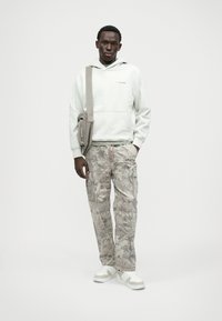 Hellgrauer Kapuzenpullover mit Kängurutasche, kombiniert mit beigen Cargo-Hosen mit einem Blattmuster. Weiße Sneakers und eine Umhängetasche.