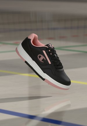Zapatilla baja negra y rosa con cordones negros, entresuela blanca y detalles en rosa, flotando sobre el suelo de una pista deportiva.