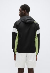 Črno-zeleni vetrovnik z nastavljivo kapuco, belimi poudarki na rokavih in elastičnim pasom, izdelan iz lahkega materiala.