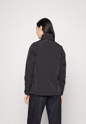 Kvinde med langt mørkt hår iført en sort letvægtsjakke og mørke bukser, stående med ryggen mod en ensfarvet hvid baggrund.