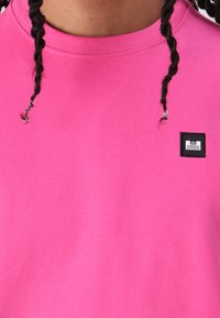 Sudadera de cuello redondo de color rosa brillante, hecha de algodón, con una etiqueta de parche negra con "WEEKEND OFFENDER" cosida. Textura suave.