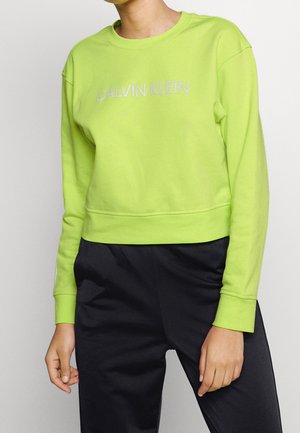 Osoba ubrana w limonkową bluzę Calvin Klein i czarne spodnie, pokazana od szyi do bioder na jednolitym tle.