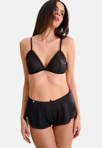 Ensemble de lingerie noire en tissu transparent. Le soutien-gorge présente une forme triangulaire avec une bordure en dentelle, associé à un short fluide avec un ourlet en forme de vague.