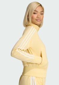 Yellow zip-up track jacket made of smooth fabric, featuring white three-stripe accents on the sleeves and a fitted waist.