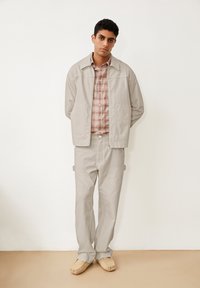 Giacca grigia chiara con colletto, abbinata a pantaloni coordinati; sotto, una camicia a quadri rosa e bianca e scarpe slip-on beige.