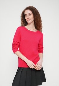 Φούξια πουλόβερ με στρογγυλή λαιμόκοψη, ριμπ μανσέτες και τελείωμα. Φορεμένο πάνω από μια μαύρη πλισέ φούστα, προβάλλοντας μια απαλή υφή.