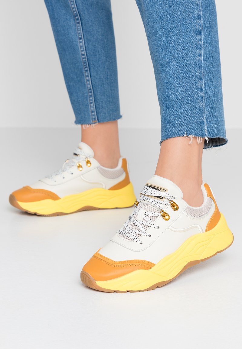 White and yellow sporty sneakers with mesh accents, textured rubber sole, and metal eyelets. Laces are white with black woven pattern.