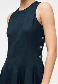 Abito senza maniche blu scuro con corpetto aderente e quattro bottoni dorati allineati verticalmente sul lato sinistro.