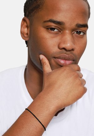Black bracelet on wrist, featuring a simple, slender design. Model has medium skin tone, short hair, wearing a white shirt.