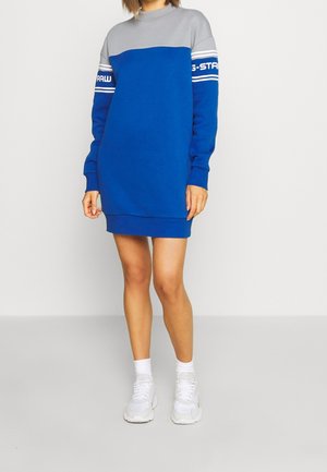 Femme portant une robe sweat-shirt bleu et gris à manches longues avec des baskets blanches, debout devant un fond clair uni.