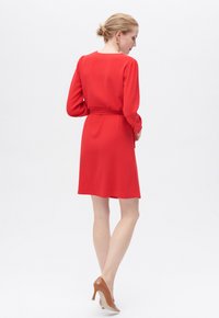 Rotes Kleid mit langen, gepufften Ärmeln und tailliertem Schnitt. Der Stoff fällt sanft, hat eine glatte Textur und einen knielangen Saum. Trägt dazu beigefarbene Absätze.