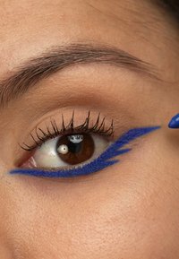 Blå eyeliner påført i en skarp vinge-design på en nærbilde av et øye, som fremhever lange vipper og en varm brun iris. Hudtonen er nøytral.