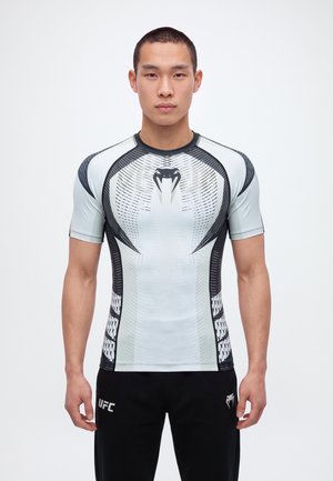 Un homme se tient face à l’objectif devant un fond uni, vêtu d’un haut de compression UFC blanc et noir et d’un short noir.
